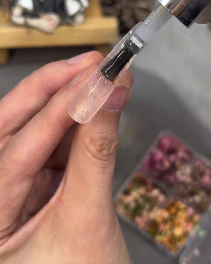 Un pincel aplicando una capa gruesa de gel transparente sobre la uña con flores y pedrería, alisando la superficie.