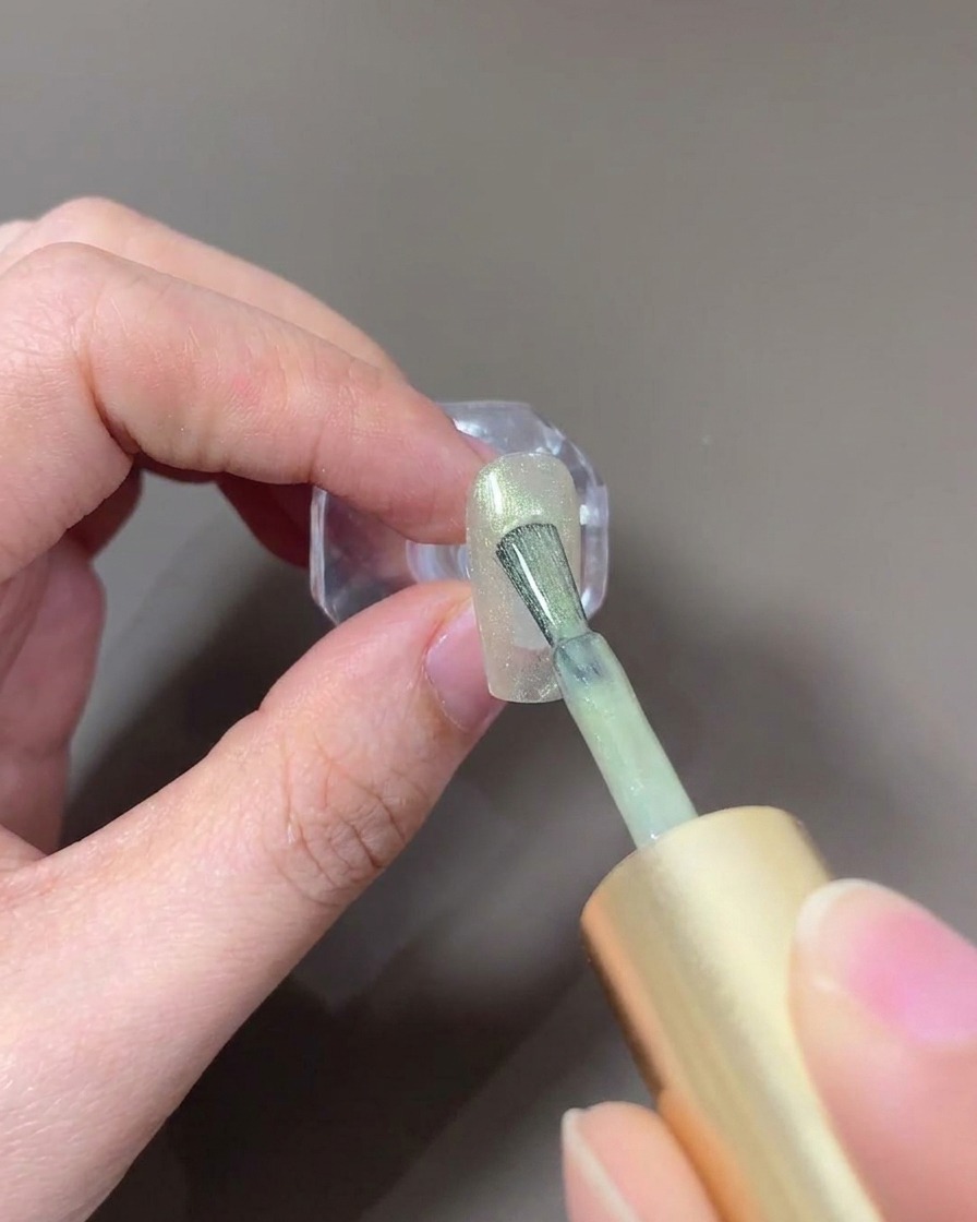 Un pincel de uñas aplicando gel ojo de gato cristal de color claro sobre una uña.