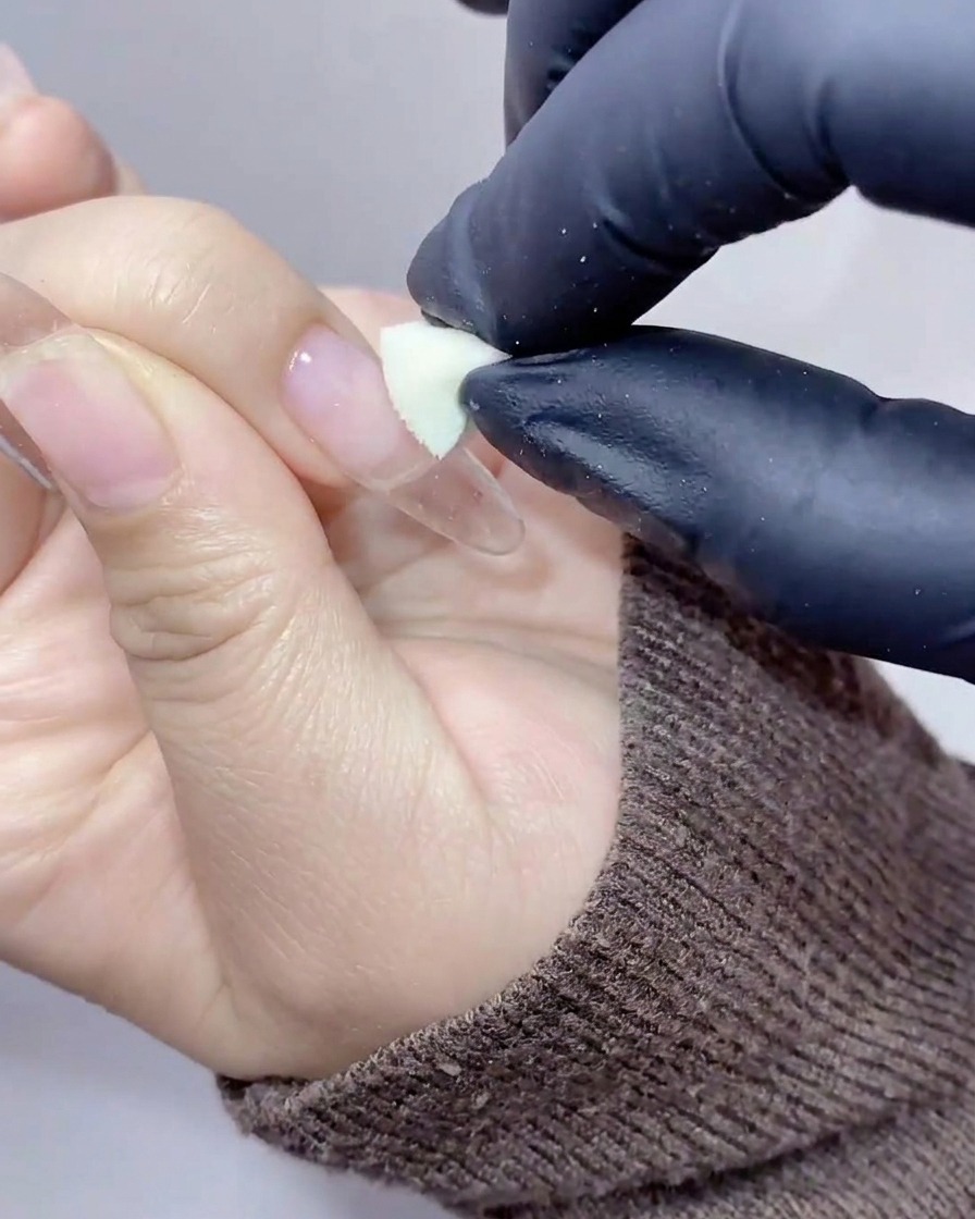 Manos enguantadas usando una pequeña cuña de esponja cosmética para aplicar gel color melocotón-naranja en el centro de una extensión de uña clara.