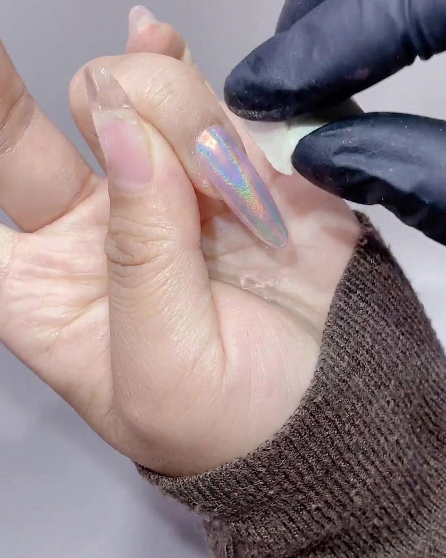 Primer plano de una mano con uñas largas siendo limpiada del exceso de polvo aurora usando una pequeña esponja de maquillaje blanca.