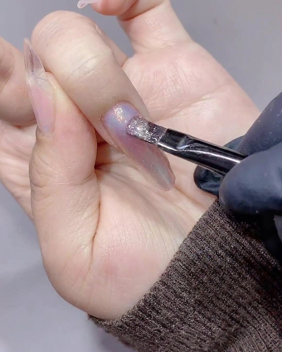 Un pincel aplicando un gel magnético cat-eye transparente y brillante sobre una uña ya decorada con polvo aurora.