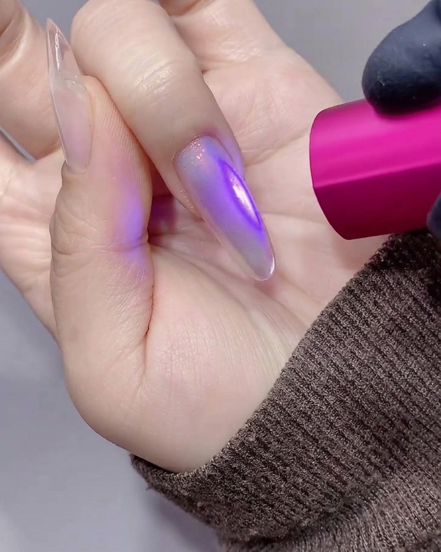Primer plano de una mano con una uña estilete translúcida con tonos púrpuras siendo curada bajo una pequeña lámpara UV/LED rosa de mano.