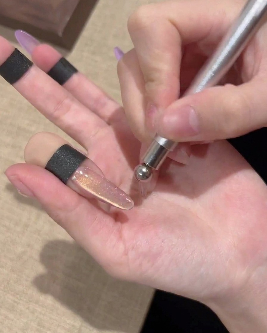La punta esférica de una varita magnética creando un patrón de remolino circular en una uña de gel con efecto ojo de gato.