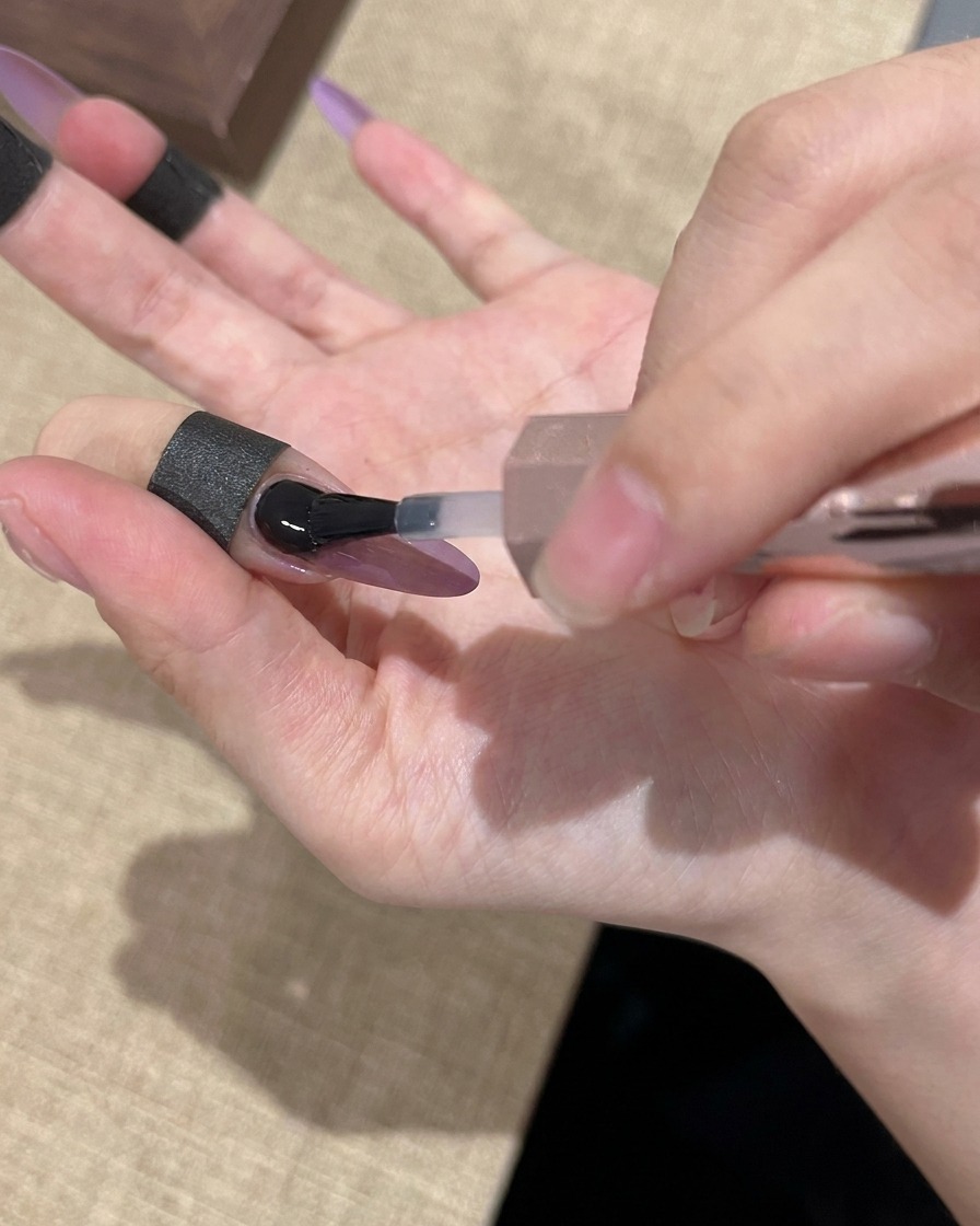 Un pincel aplicando un top coat transparente y brillante sobre una uña con diseño ojo de gato morado y dorado.