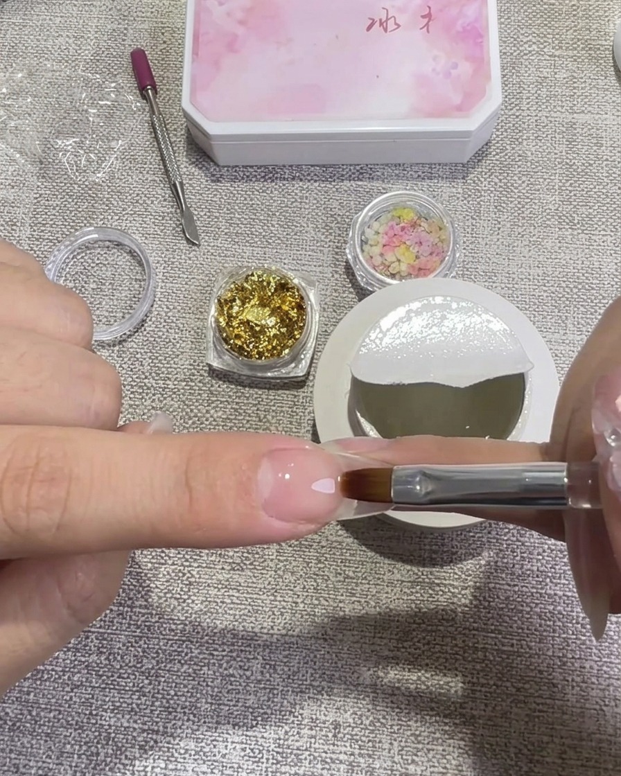 Una mano utilizando un pincel plano para aplicar gel rígido transparente sobre una uña, con frascos de lámina de oro y flores secas de fondo.