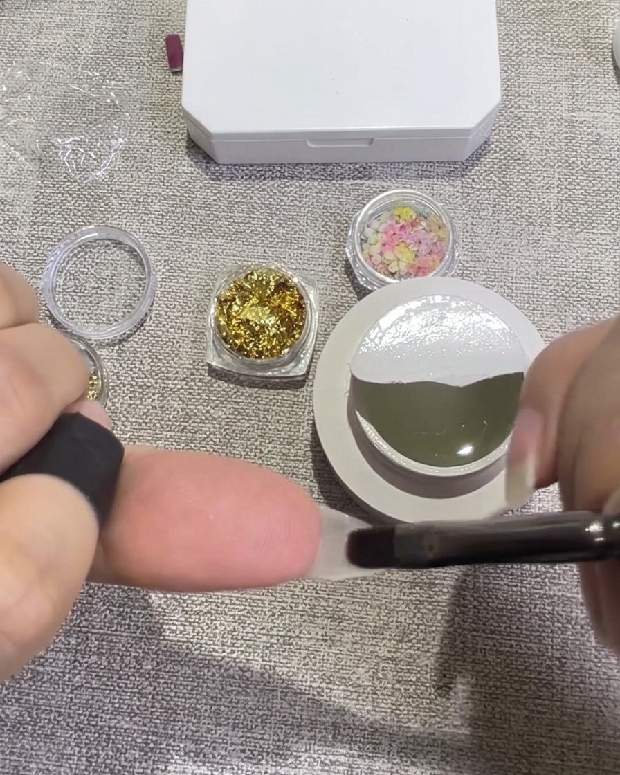 Pinzas colocando pétalos de flores secas de colores sobre una uña cubierta con gel transparente húmedo.