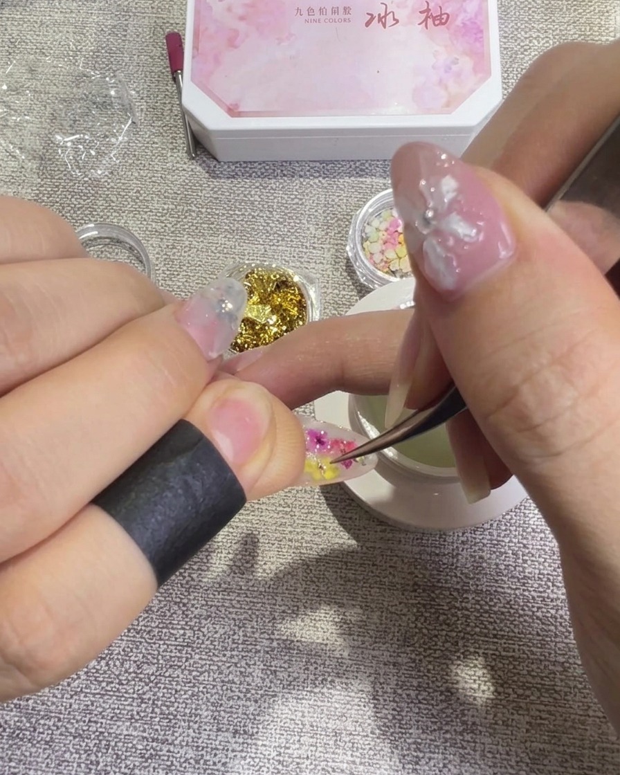 Pinzas ajustando delicadamente un pétalo de flor seca en la superficie de la uña para lograr un diseño equilibrado.
