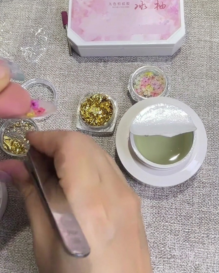 Pequeños copos de lámina de oro siendo aplicados sobre la superficie de la uña cerca de las flores con pinzas.