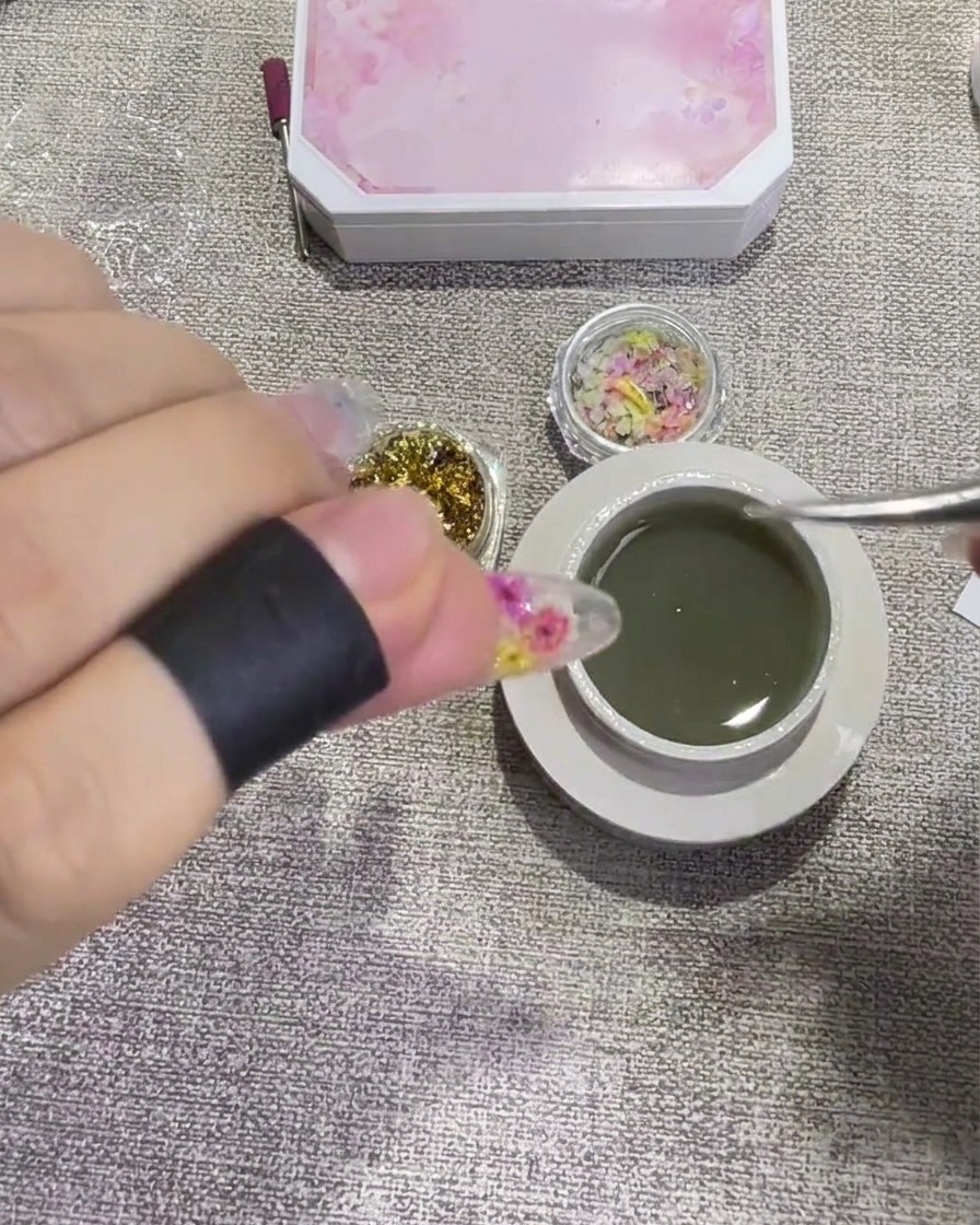 Primer plano de unas pinzas colocando una pequeña pieza brillante de lámina de oro sobre una uña con gel.