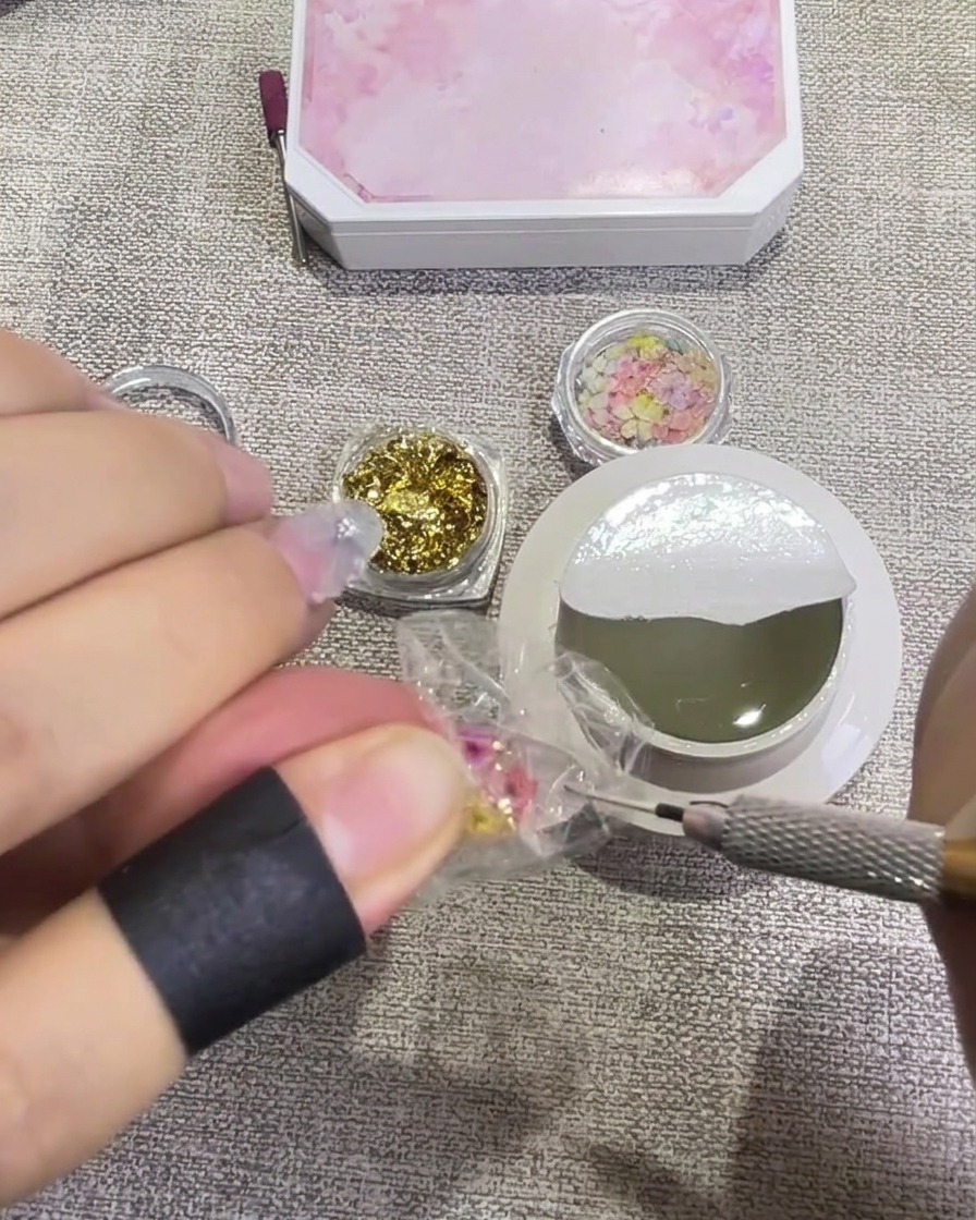 Un pincel aplicando un top coat brillante transparente sobre una uña decorada con flores y oro, dándole un aspecto húmedo y translúcido.