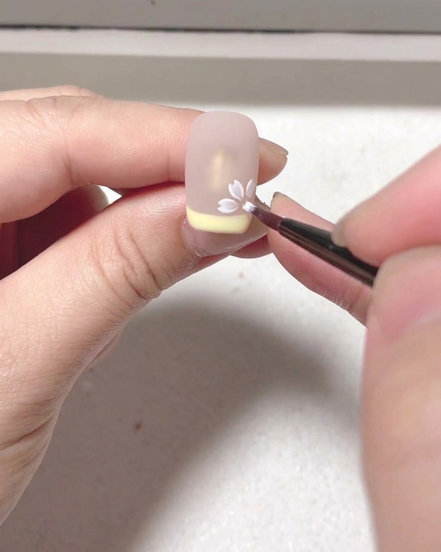 Una vista en primer plano de un pincel aplicando blooming gel blanco para crear la forma de un pétalo sobre una superficie de uña nude.