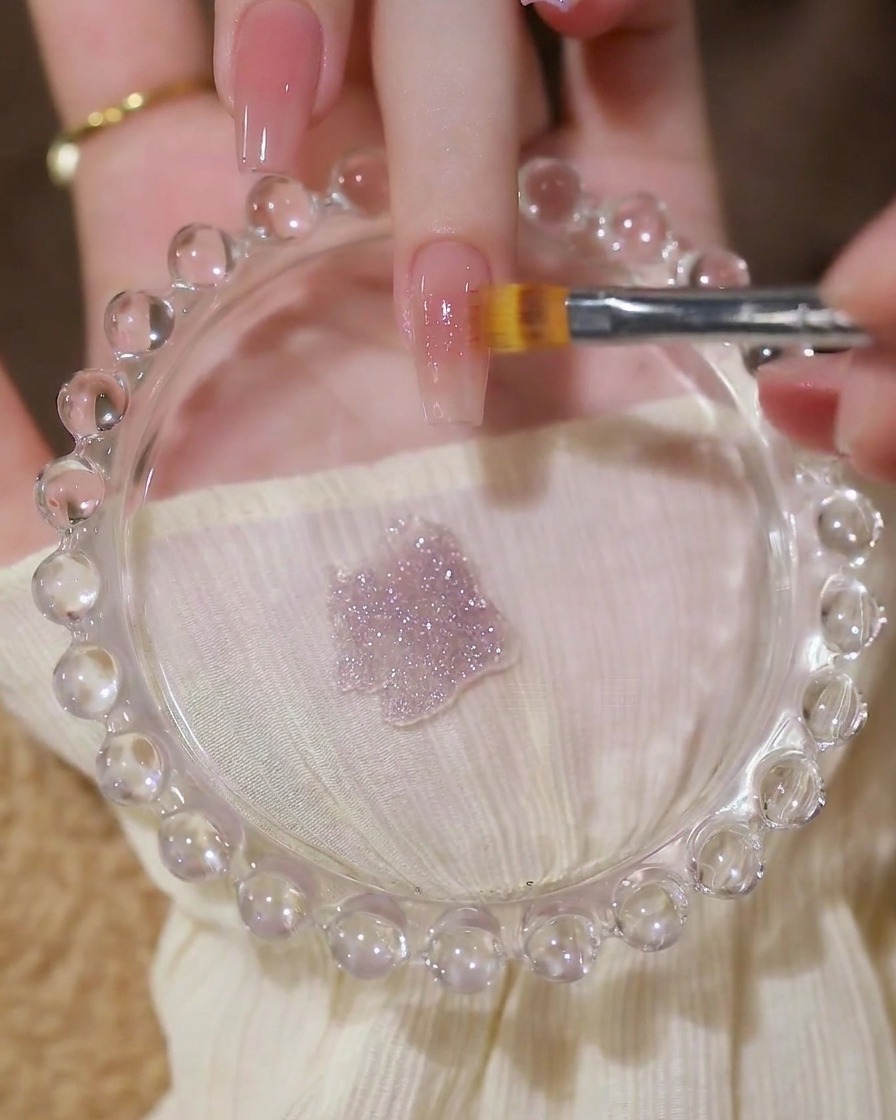 Un pincel aplicando una capa de gel de lentejuelas transparente y brillante sobre una base de uña nude.