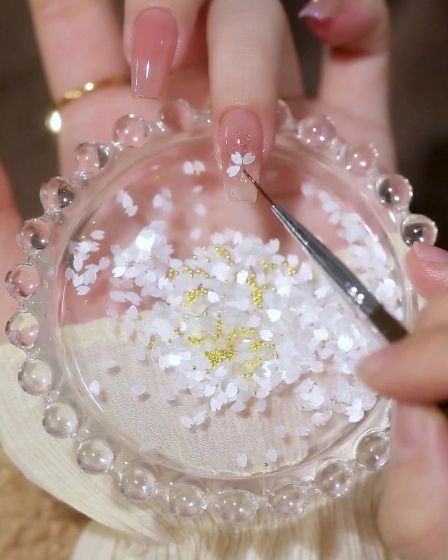 Un primer plano de un pincel de detalle colocando lentejuelas en forma de pétalos blancos sobre una uña en un patrón de flores.