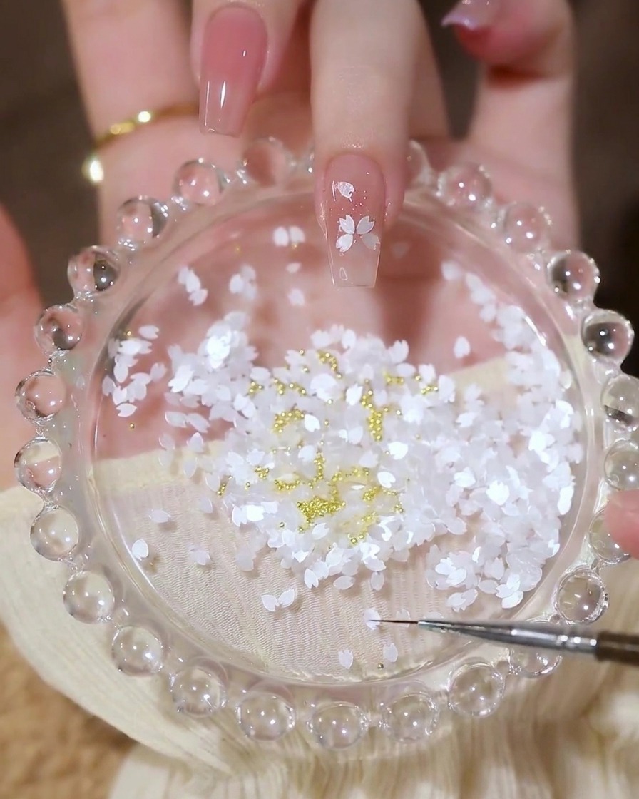 Una uña con un diseño de flor completado hecho de lentejuelas de pétalos, con una pequeña cuenta dorada en el centro.