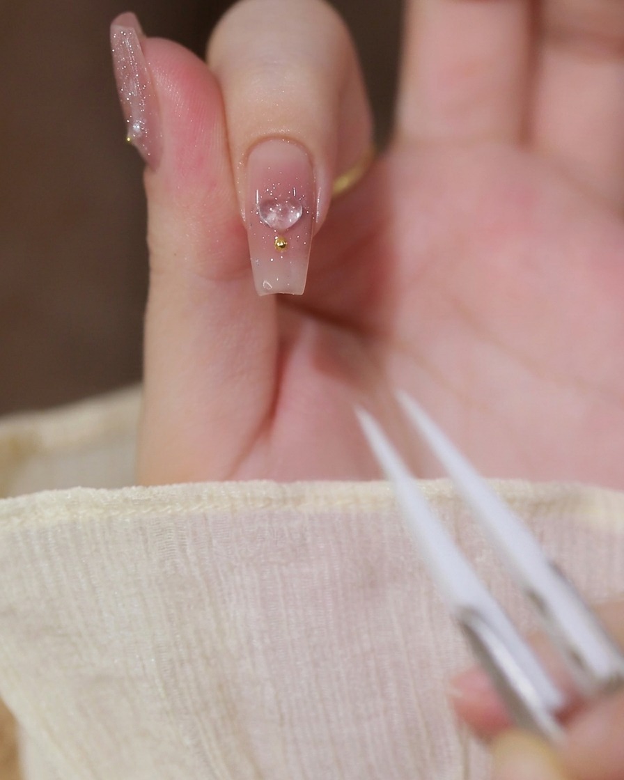 Un primer plano de un dedo índice con una base de gel nude lechoso, con un dije de corazón 3D transparente y una cuenta dorada, con pinzas visibles en primer plano.