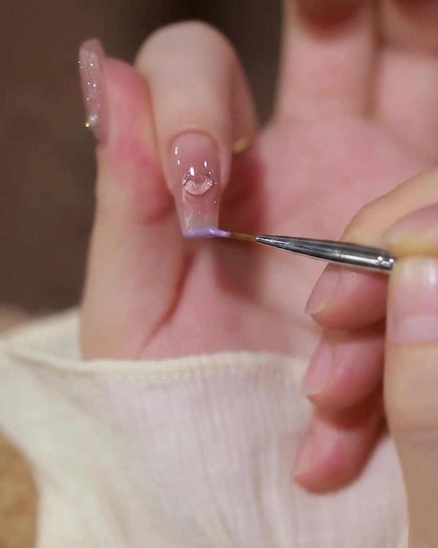 Un primer plano de un pincel delineador fino aplicando esmalte de gel morado pastel a lo largo del borde de un dedo índice con un dije de corazón.