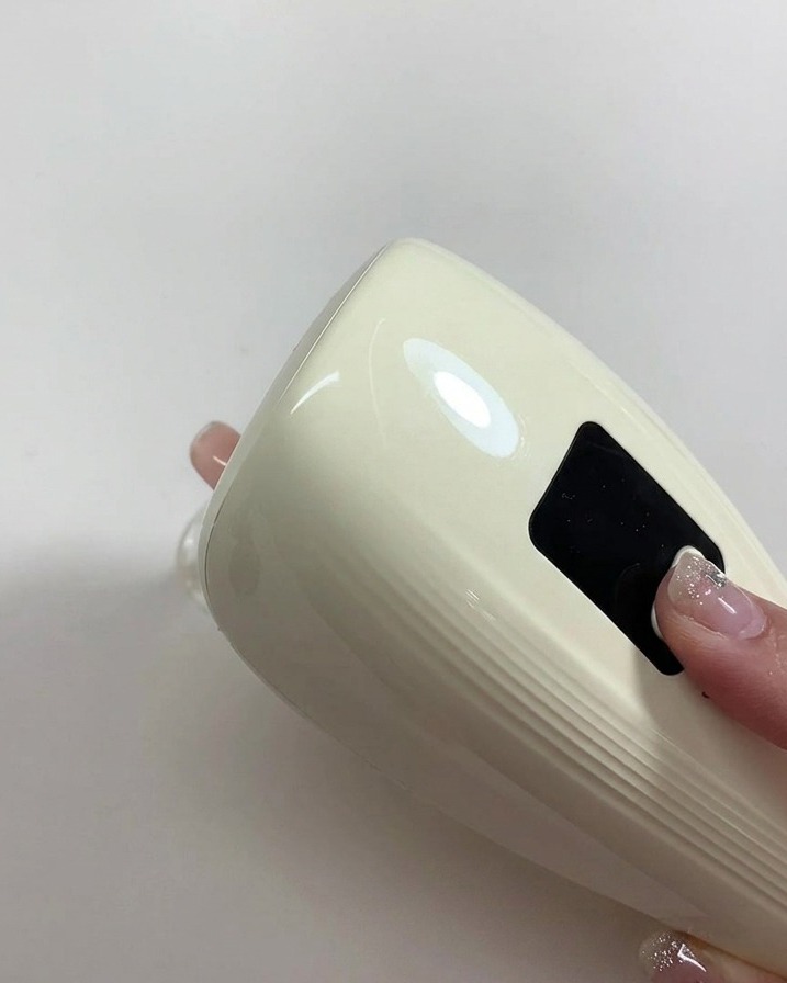 Una mano sosteniendo una lámpara UV/LED portátil sobre una uña recién pintada de color nude.