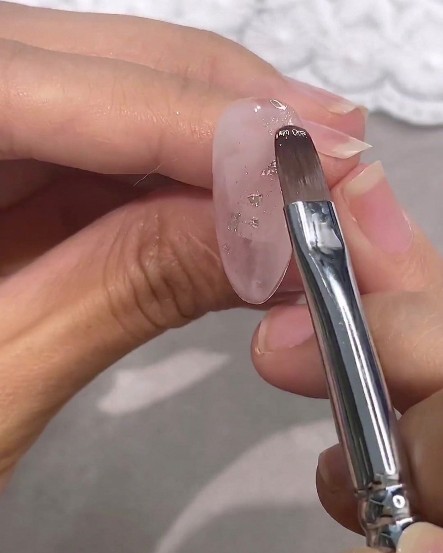 Un pincel aplicando un esmalte rosa transparente sobre una uña con escamas plateadas.