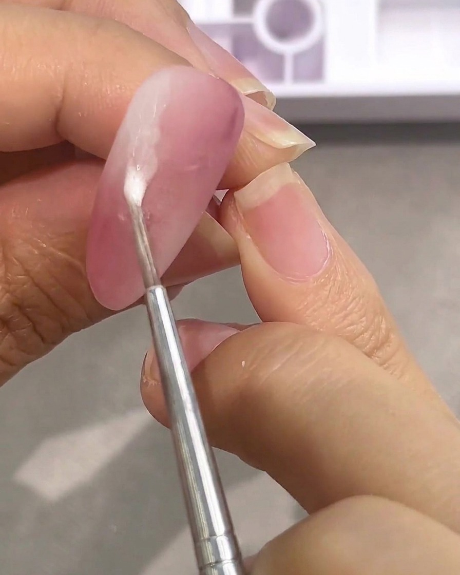 Un pincel aplicando gotas de pintura blanca sobre una uña rosa mate.