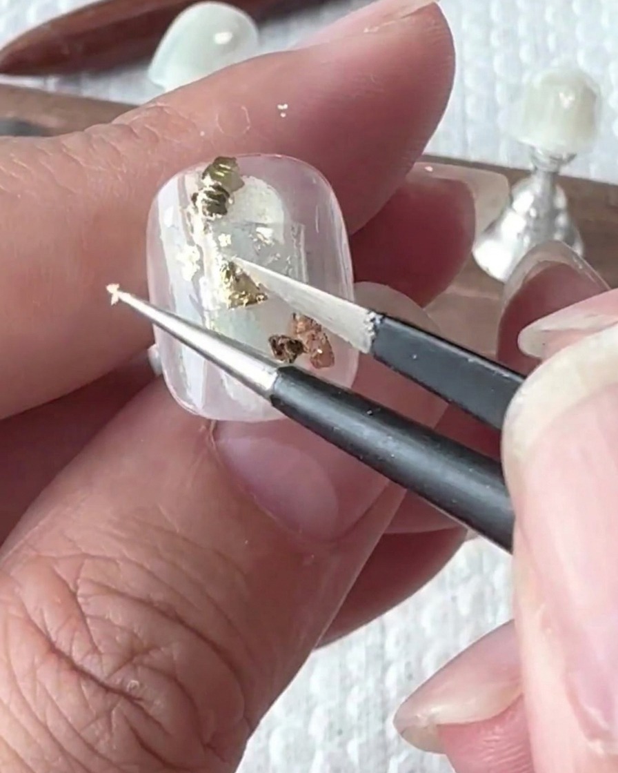 Unas pinzas colocando cuidadosamente trozos irregulares de pan de oro sobre la superficie de la uña.