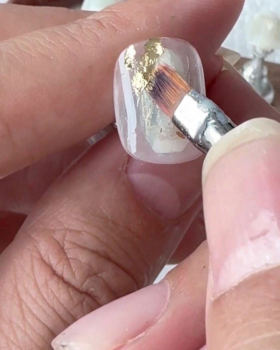 Primer plano de una herramienta de silicona presionando los copos de pan de oro aplicados a una uña.