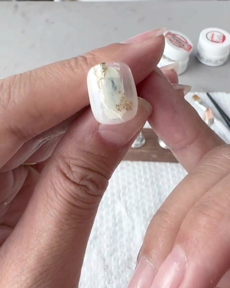 Primer plano de un pincel plano mezclando y difuminando delicadamente las líneas de esmalte en gel blanco en la punta de una uña.