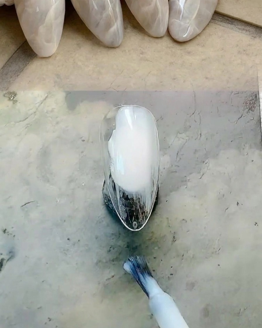 Un pincel de botella aplicando esmalte de gel blanco lechoso sobre la superficie de la uña.