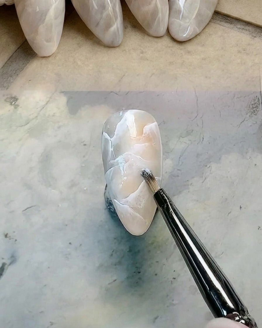 Un pincel de detalles agregando y difuminando gel blanco adicional sobre un diseño de uña marmoleada.