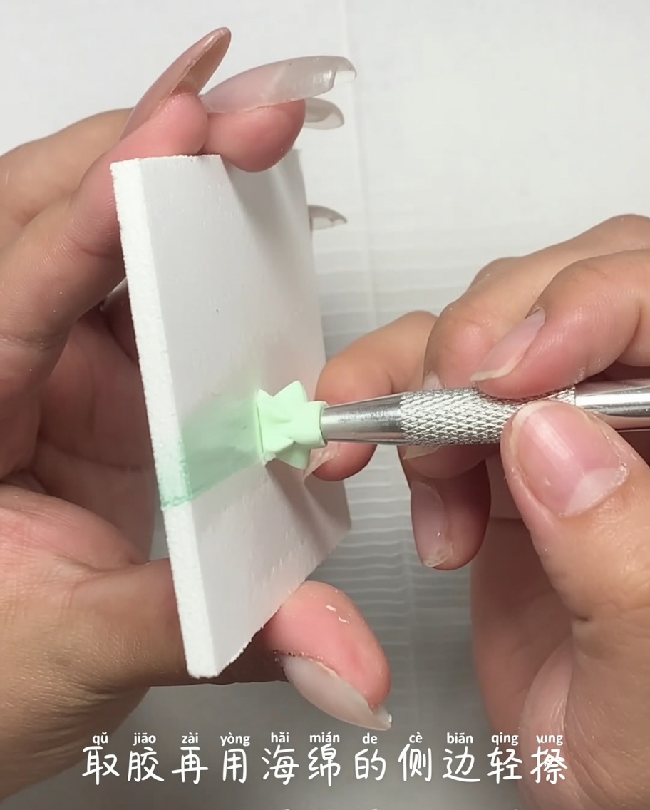 Vista de cerca de una herramienta metálica aplicando una pequeña cantidad de esmalte de gel verde claro desde una paleta hacia el borde de una esponja de maquillaje.