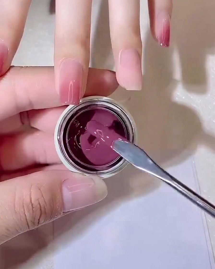 Un palito mezclador de metal levantando una pequeña gota de gel jelly color púrpura-rosa de un frasco pequeño.
