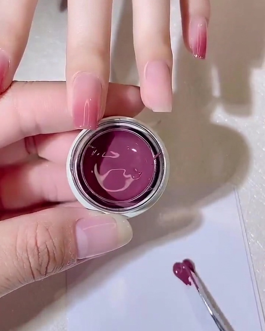 Una herramienta de metal extendiendo una pequeña cantidad de gel púrpura-rosa finamente sobre un papel de paleta blanco.