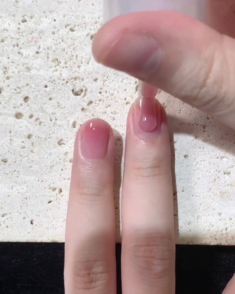 Primer plano de un pincel aplicando esmalte jelly rosa traslúcido cerca del área de la cutícula de una uña natural