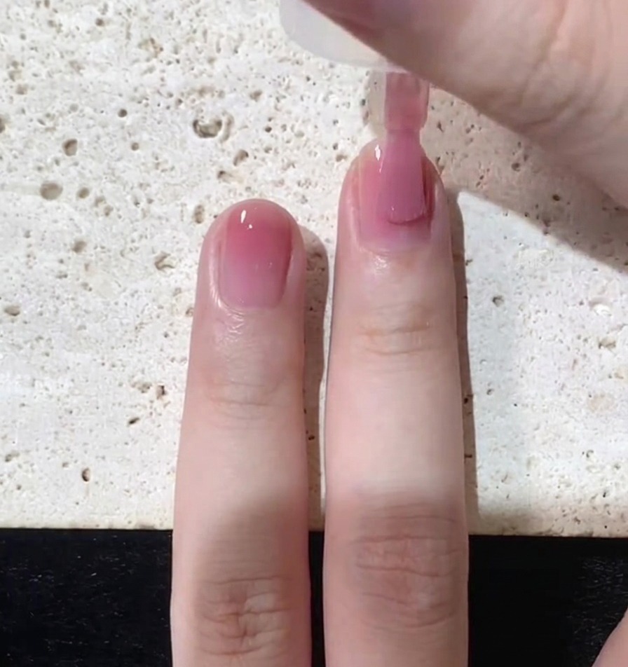 Primer plano de un pincel aplicando una capa fina de esmalte rosa traslúcido sobre una uña natural