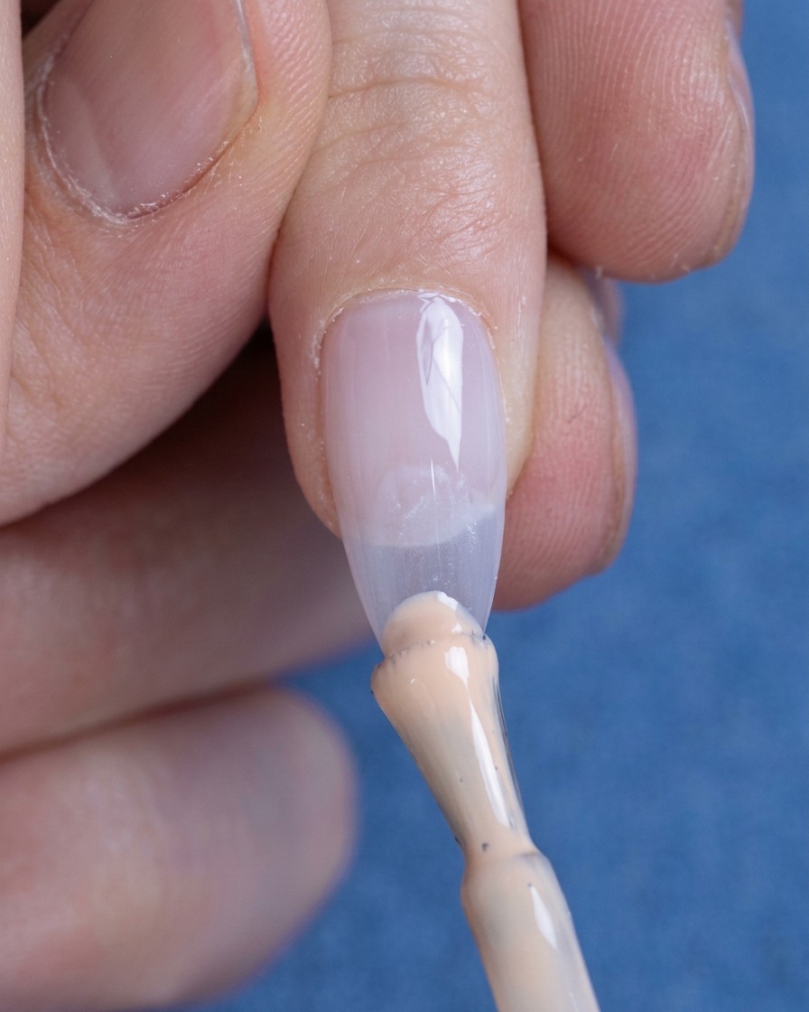 Una pequeña gota de esmalte de gel nude siendo aplicada en la punta de la uña.