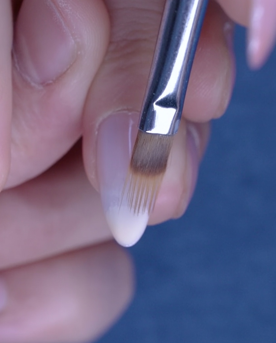 Un pincel plano difuminando el esmalte de gel nude hacia arriba en la uña para crear un efecto ombré.