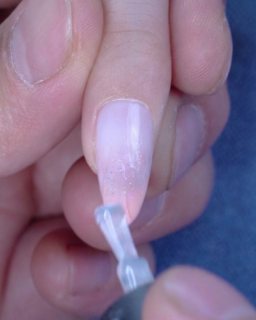 Un pincel aplicando una capa suave y transparente de top coat brillante sobre una uña ombré con glitter.