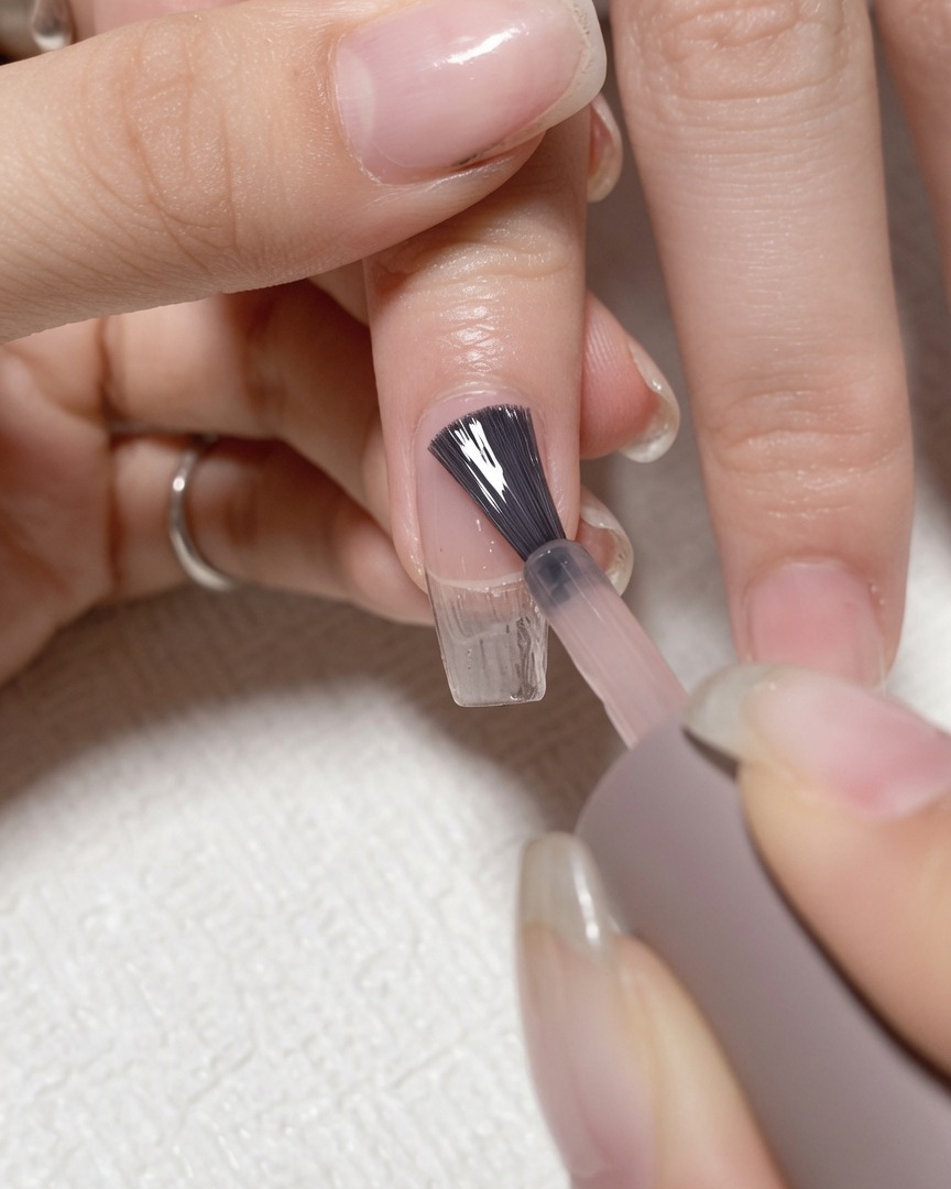 Un pincel aplicando gel base transparente sobre una uña natural limpia.