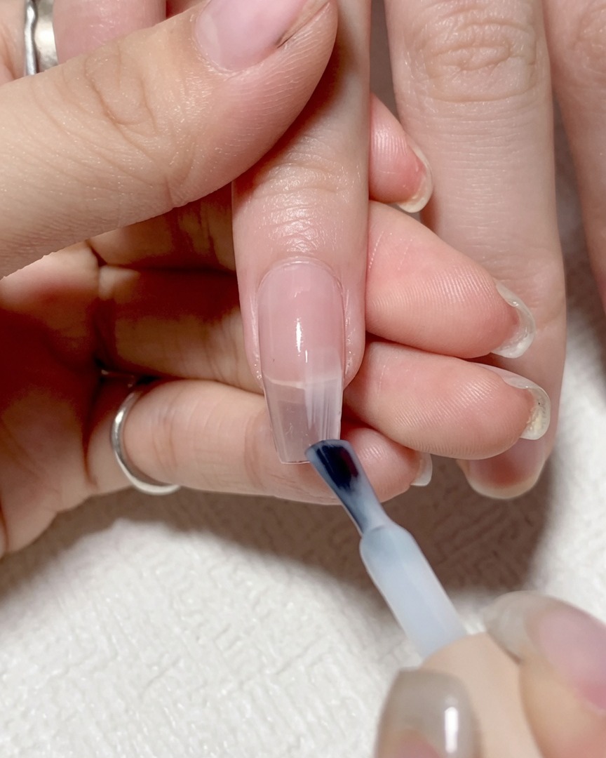 Un pincel de uñas aplicando un gel transparente de tono claro en la mitad derecha de la uña.