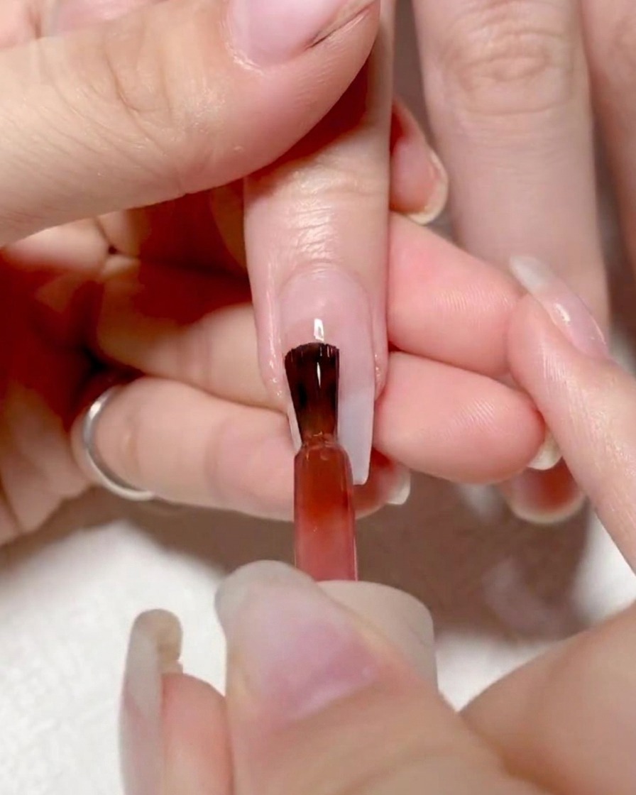 Un pincel de uñas aplicando un gel rojo vibrante en la mitad izquierda de una uña.