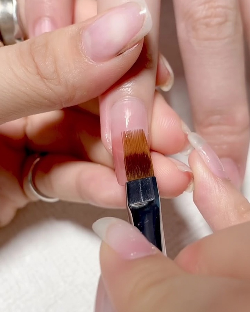 Un pincel siendo arrastrado suavemente hacia abajo sobre la superficie de la uña para mezclar los colores en un ombré suave.