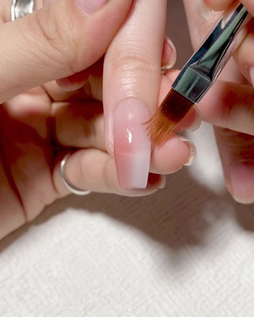 Un pincel aplicando un gel fortalecedor transparente y ligeramente espeso sobre toda la superficie de la uña.
