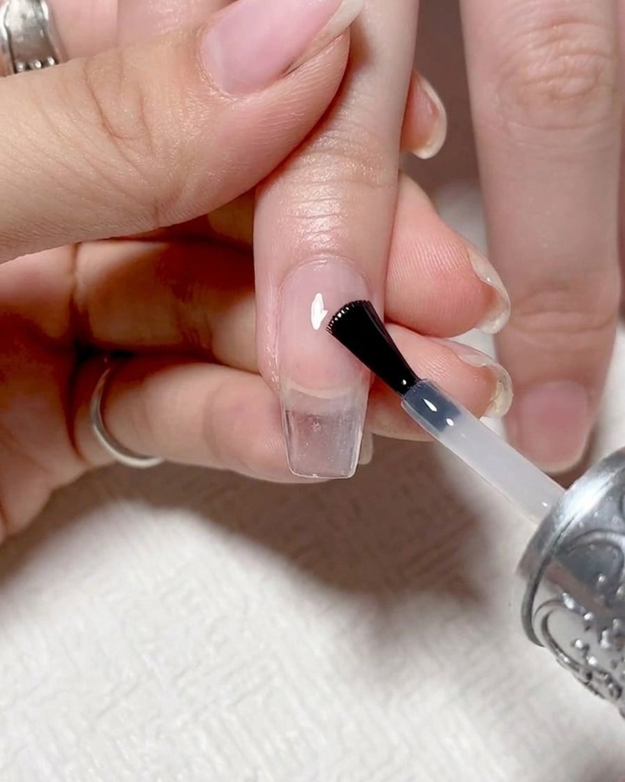 Primer plano de un pincel de uñas aplicando un top coat transparente y brillante sobre una uña con degradado terminada.