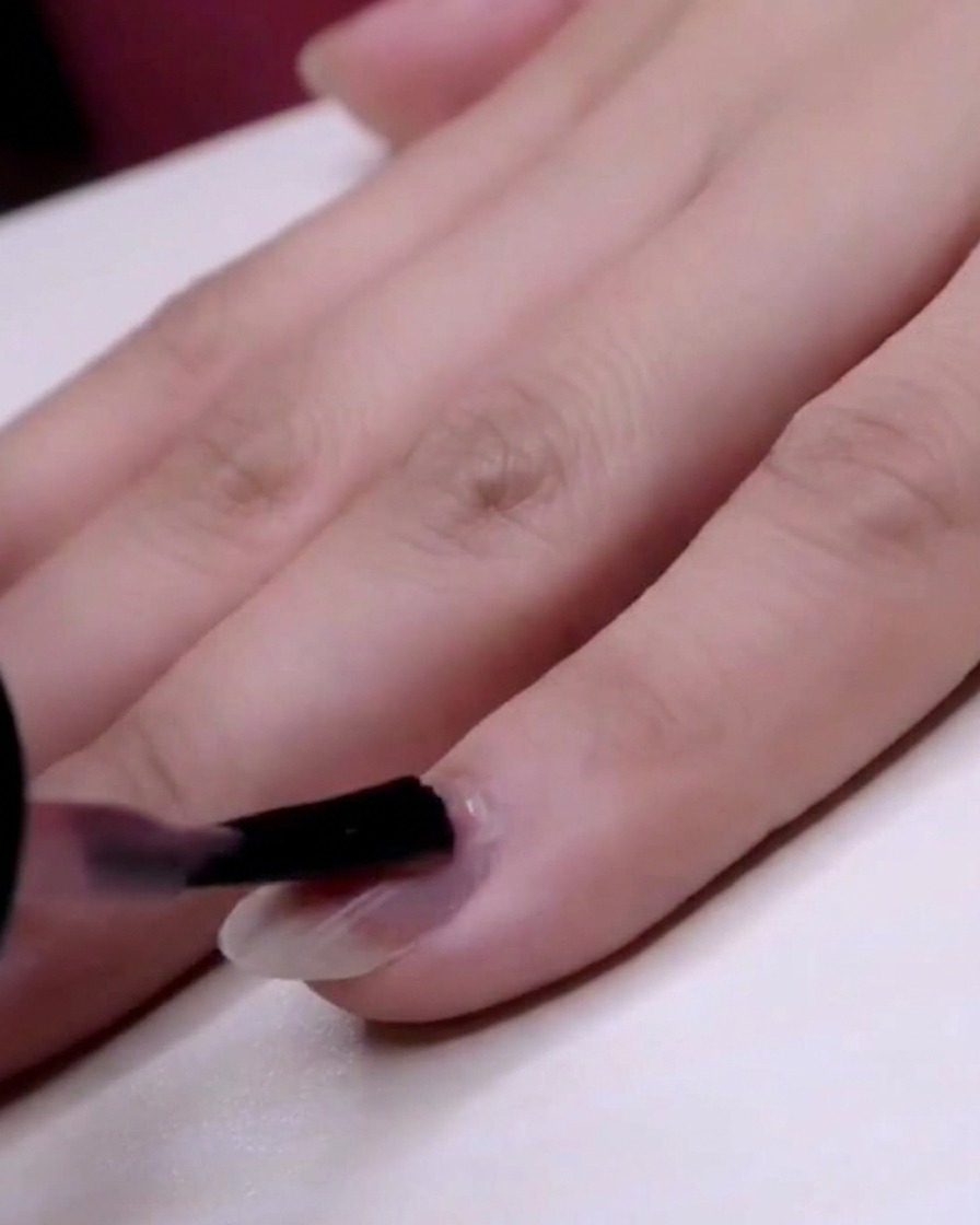 Un pincel aplicando esmalte rosa jelly transparente sobre toda la extensión de la uña del dedo meñique.