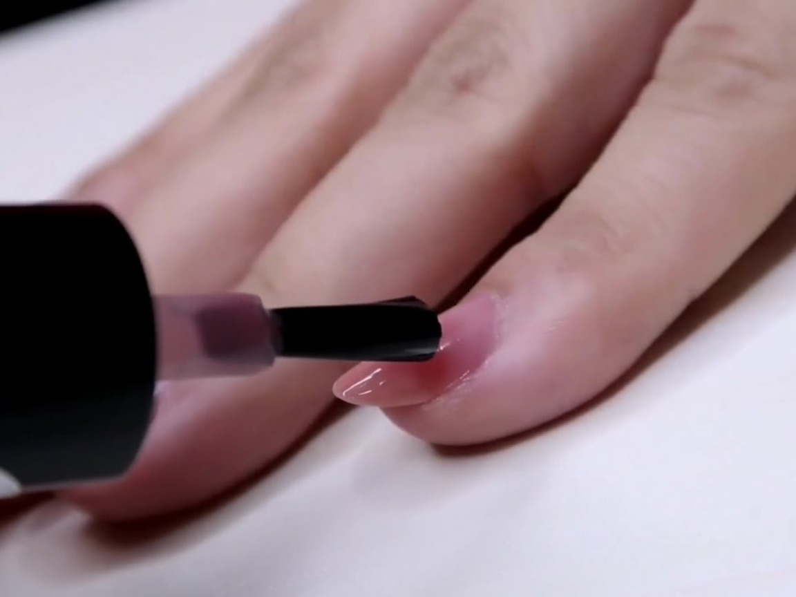 Un pincel aplicando una capa de esmalte rosa transparente a la mitad exterior de una uña.