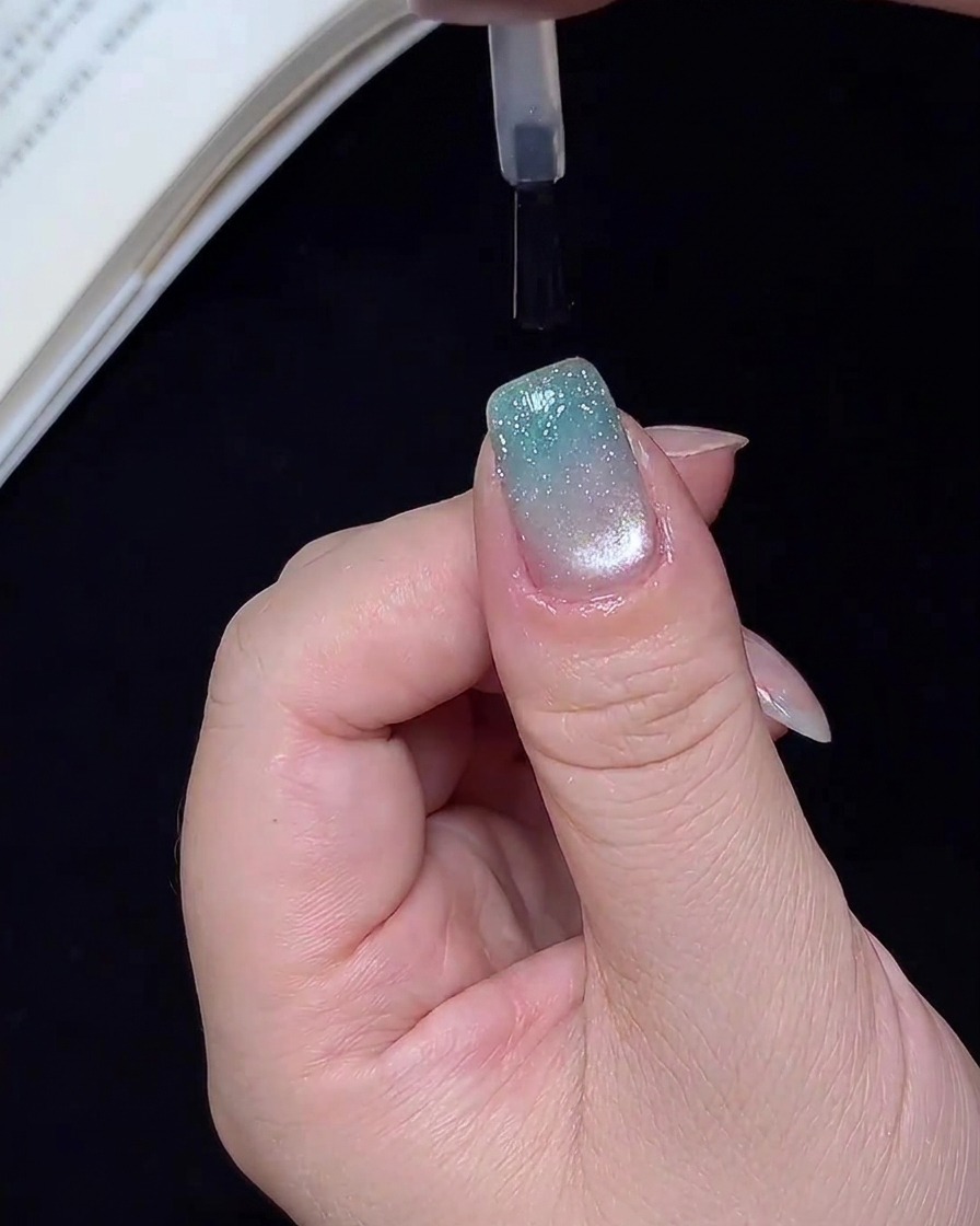 Un pincel aplicando una capa superior transparente y brillante sobre una uña terminada con degradado de purpurina azul.