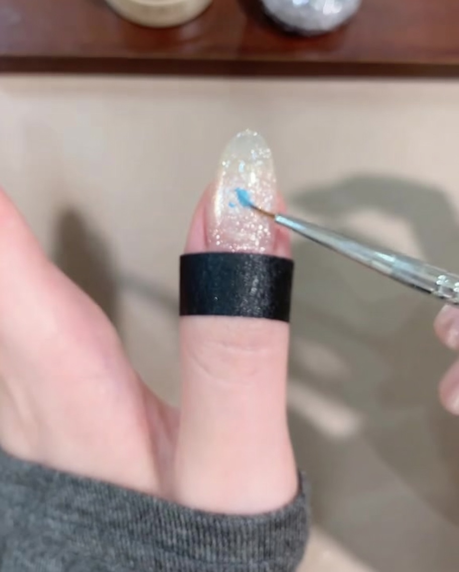 Un pincel liner fino aplicando una gota pequeña de gel azul en el centro de la uña.
