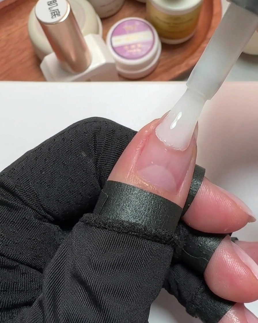 Primer plano de un pincel aplicando esmalte de gel blanco lechoso sobre la uña del pulgar.