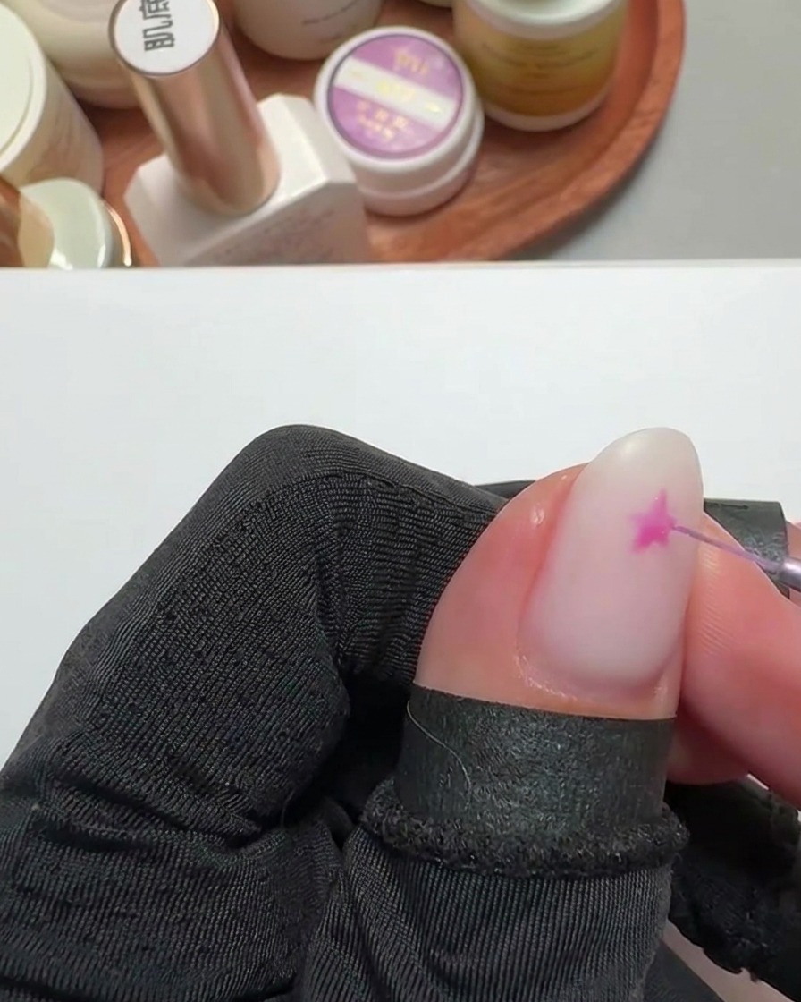 Primer plano de un pincel liner fino pintando una estrella rosa brillante sobre una uña con base blanco lechoso.