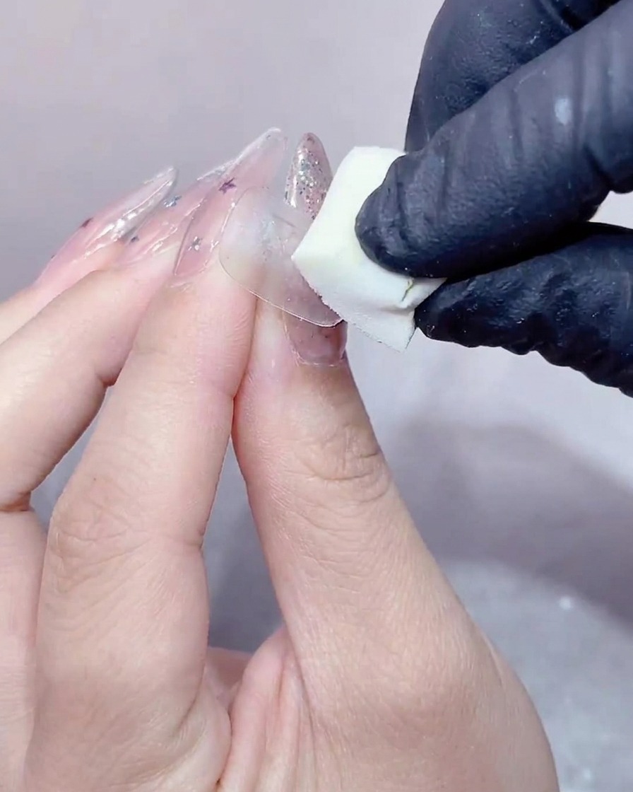 Una mano con guantes presionando una pequeña esponja blanca de maquillaje sobre la superficie de una uña cubierta con gel transparente.
