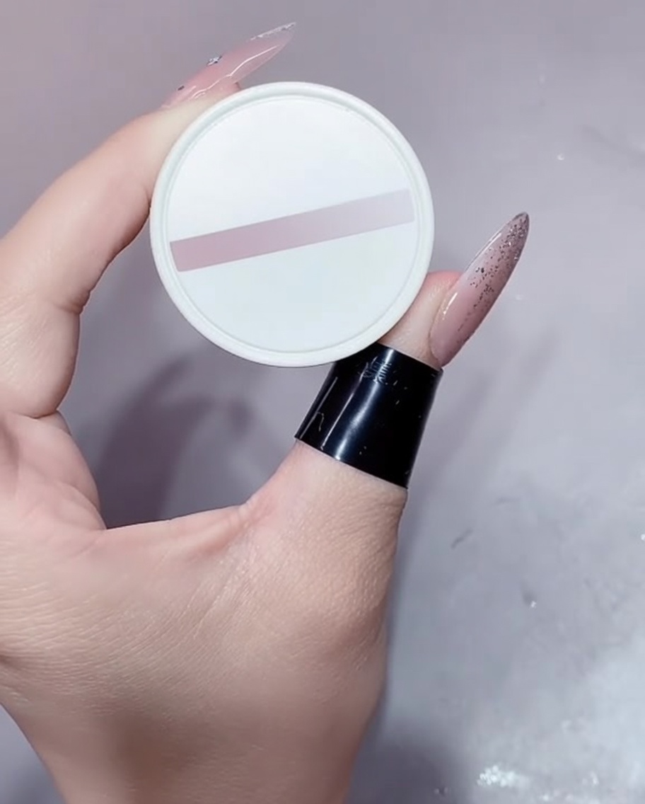 Un pequeño bote blanco de esmalte en gel color nude sostenido por una mano con la manicura hecha.